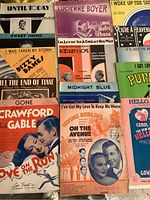 Top view of various musical score sheets and songbooks laid out; shows colorful vintage covers with titles including 'Love on the Run', 'Until Today', and 'I've Got My Love To Keep Me Warm'.