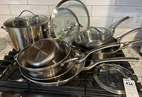 Wide shot showing entire collection of stacked Calphalon pots, pans, and lids with metal handles and glass tops on stove.