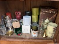 Photo showing assortment of new boxed and packaged candles and various candle holders on wooden shelf.