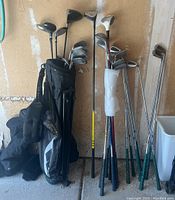 All golf clubs and golf bag shown standing against wall with some club head covers visible
