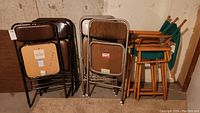 Two sets of folding chairs: brown metal Samsonite folding chairs and wooden wooden directors chairs with green canvas seats and backs, stacked in basement corner