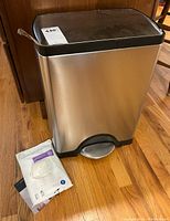 Simple Human stainless steel trashcan with a black lid and foot pedal on a hardwood floor, accompanied by unopened trashcan bags in packaging.