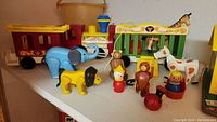 Close-up photo showing vintage Fisher Price toys including plastic and wood animal figures (elephant, lion, horse, cow), a small circus cage play set, and character figures like a clown and monkey.
