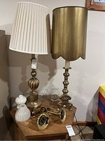 Photo showing two brass table lamps, one with pleated white fabric shade and the other with scalloped brass shade, along with various small glass and ceramic items on a wooden table.