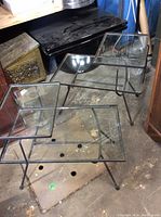 Two glass and metal end tables with rectangular tops and lower glass shelves on metal tubular frames, positioned in a storage area.