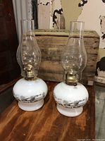 Pair of white glass oil lamps with clear chimneys and brass burners shown from front and side on wooden surface.