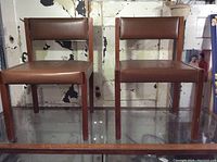 Two oak framed chairs with brown vinyl upholstery side by side, showing design and condition.