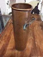 Photo of the copper vase showing its cylindrical shape, dual side handles, and aged copper surface with patina.