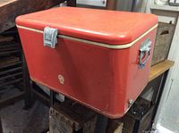 Red metal cooler with metal hinges, clasp, and handles, placed on a wooden surface.