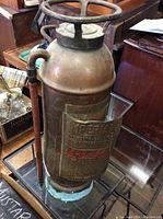 Full view of the vintage copper fire extinguisher showing the cylindrical tank, top handle, and attached piping on a base stand.