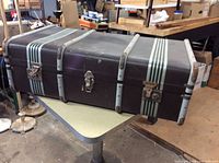 Top and side view of large brown vintage trunk with two white stripes running lengthwise, metal corner reinforcements, and three metal clasp closures including central lock.