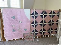 Both large Amish quilts displayed on a wooden rack with the small pillow placed on the floor against the rack. One quilt is pink with floral appliques, the other has a geometric pink and black diamond pattern.