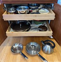 Cabinet showing two wooden pull-out drawers containing multiple saucepans, frypans, and cake molds, along with items placed on wooden floor including two frypans and one teapot