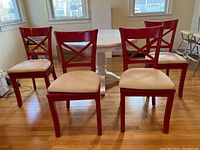 Front view of four red wooden dining chairs with beige cushions around a white table in a room with wooden floor and windows.