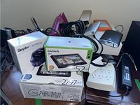 Overview of mix of electronic devices mainly power chargers, GPS units in box, power adapters, and power strips on a table.