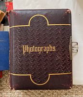 Cover of antique photo album with 'Photographs' embossed in gold on dark brown leather cover with embossed basket weave pattern.
