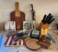 Overview of all items in the lot on a kitchen counter including knives in block, cutting boards, ceramic trivets, cake cutters, and coasters.