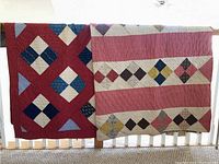 Two patchwork quilts hanging side by side over a railing, showing their full patterned surfaces and colors including reds, pinks, blues, and creams.