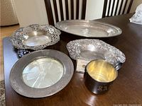 Five silver items and coin displayed on table showing cup, wine coaster, note chip, two decorative platters, and oval tray