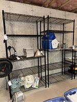 Two black wire metal shelving units against a basement concrete wall, with various household items on the shelves including fans, lamps, a cooler, glass jars, papers, and a blue bag. The shelving units have five wire shelves each.