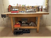 Front view of wood work bench showing attached metal vice on left side, hand tools mounted on backboard, and toolboxes on bench and floor.