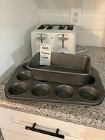 Photo of white four-slice toaster, metal loaf pan, and metal muffin pan with 12 cups on countertop