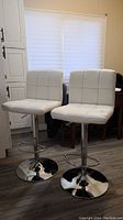 Pair of white vinyl cushioned adjustable bar stools with tall backs, chrome round bases and footrests, placed on wood floor.