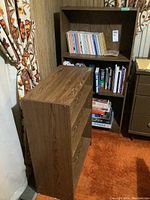 Tall laminate bookcase filled with books and a small glass dish on top shelf, shown with curtains and adjacent furniture.