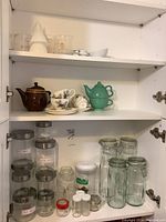 Glass jars, canisters, ceramic teapots and teacups inside kitchen cabinet.