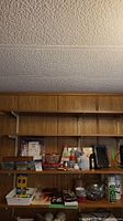 Wide view of two wooden shelves containing assorted crafting supplies including glue, tape, paint, brushes, marbles and other items.