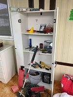 Front view of cupboard with door open, showing four shelves containing cleaning and hand tools, a mop, and various small items.