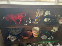 Shelf with bright red bull figurine, white ceramic miniature tea cups and saucers, small figurines, blue and white Delft style plate, wooden bowls, large floral soup mugs, black ceramic vase, and assorted figures.
