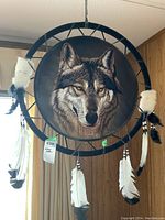 Front view of signed dream catcher displaying a realistic wolf face painting in the center with hanging white feathers tipped in black and fur pieces.