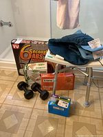 Photo showing boxed 6 Second Abs exercise machine, black dumbbells, shower stool, Solaray heating pad, muscle pad, and digital blood pressure monitor on a tiled floor and shower stool.