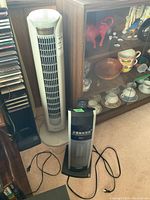 Two fan/heaters placed on the floor near a cabinet and a collection of cups and saucers. One is a tall white Tower Airtech tower fan/heater, the other is a smaller black and silver Bionaire unit with a remote on top.