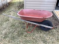 Full view of pink plastic wheelbarrow with single rubber wheel and metal frame with wooden handles showing outdoor grass and siding background