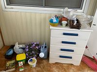 Wide view of the gardening supplies lot showing the watering can, pots, planter trays, Miracle-Gro container, and gardening book on and around a white dresser.