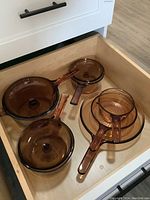 Overview showing all cookware included in the set inside a wooden drawer. Two lidded saucepans and two frying pans with handles in amber-colored translucent glass.