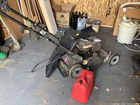 Full view of the Murray lawn mower with attached black grass collection bag and red jerry can on the concrete floor in a shed.