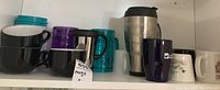 Shelf view showing assorted ceramic mugs in black, white with botanical print, and travel mugs in blue plastic, purple plastic, and stainless steel insulated mug.