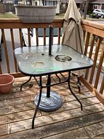 Square glass top patio table with metal frame, umbrella hole and weighted base stand, showing wear on glass top