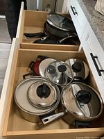Photo showing several stainless steel pots with lids in a drawer, including large and medium sizes with black handles and glass lids.