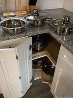 Several Apenhof brand stainless steel cookware items placed on kitchen countertop and inside cabinet corner, showing pots, pans, lids, and a bowl.