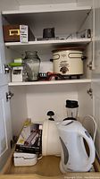 Cabinet interior showing the crockpot, glass pitcher, salad spinner, power chopper, and sharpener on shelves along with electric kettle and other items below.