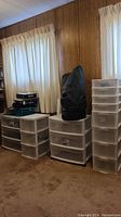 Four resin drawer storage systems on wheels placed in room along wall with carpet and curtains. Canvas bag on top of middle storage unit.