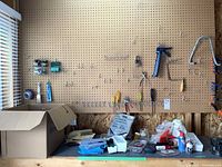 Wide shot showing pegboard with hanging screwdrivers, clips and various tools above workbench with loose items and boxes under window.