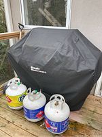 Ultra Chef BBQ grill covered with black 'Simple Measures' branded cover. Four gas cylinder tanks in front, three on the deck and one attached to the grill's side tray.