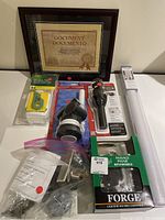 Photo displaying various household items including a wood framed picture frame in the back, door handles and lock sets in packaging, plumbing parts, and a window film kit with tools and roll of tint film arranged on a surface