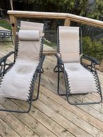 Pair of beige anti-gravity lounge chairs on wooden deck. Shows full view of chairs with fabric seats and attached pillows.
