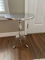 Full view of the metal silver pedestal stand showing round top, central turned column, and tripod legs on wood floor in natural light.